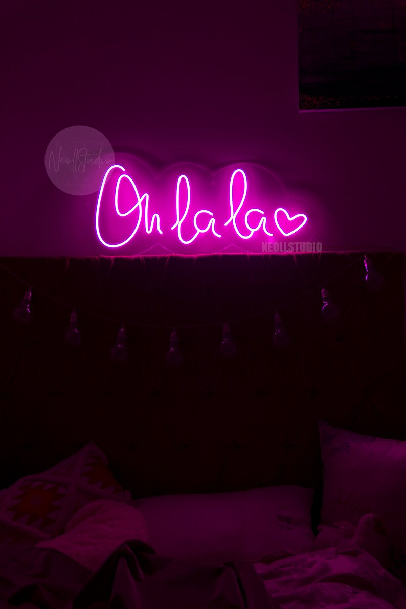 May include: A bright pink neon sign mounted on a wall, displaying the cursive text Oh la la followed by a heart symbol. The sign emits a vibrant glow, illuminating the dark space below where bedding and a string of small light bulbs are visible.
