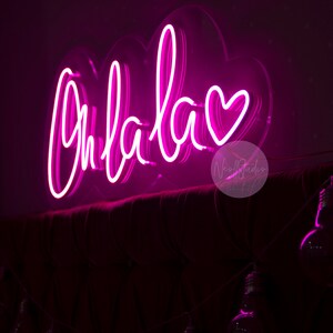May include: A bright pink neon sign featuring the cursive text Ohlala followed by a heart symbol. The sign is mounted on a clear acrylic backing shaped like a cloud and is positioned above a dark, tufted headboard with a string of light bulbs.