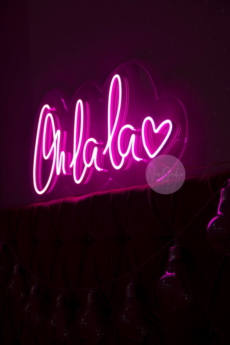 May include: A bright pink neon sign featuring the cursive text Ohlala followed by a heart symbol. The sign is mounted on a clear acrylic backing shaped like a cloud and is positioned above a dark, tufted headboard with a string of light bulbs.