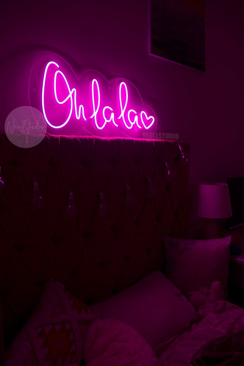 May include: A bright pink neon sign mounted on a wall, displaying the cursive text Oh la la followed by a small heart symbol. The sign is constructed with a clear acrylic backing and emits a vibrant, glowing light that illuminates the room.