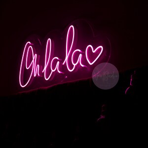 May include: A bright pink neon sign mounted on a dark surface, displaying the cursive text Oh la la followed by a heart symbol. The glowing light creates a vibrant magenta illumination against the surrounding black background, highlighting the script.