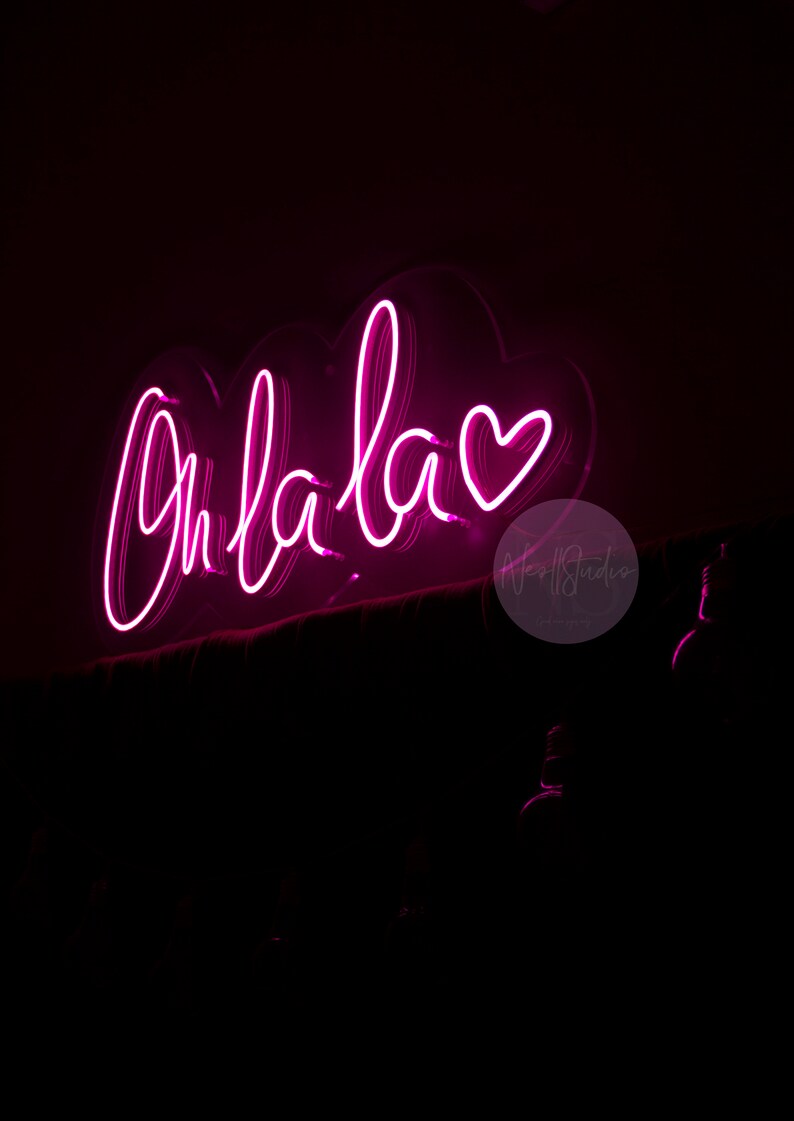 May include: A bright pink neon sign mounted on a dark surface, displaying the cursive text Oh la la followed by a heart symbol. The glowing light creates a vibrant magenta illumination against the surrounding black background, highlighting the script.