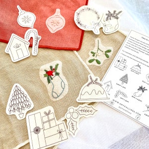 May include: A set of printable Christmas embroidery patterns featuring a variety of festive designs, including ornaments, a stocking, a Christmas tree, a house, a candy cane, a wreath, mistletoe, a pudding, and a gift box. The patterns are printed on white paper and include instructions for stitching. The text on the paper reads "How to Create", "These designs are pre-printed", "making it easier than ever to", "Simply choose your design, peel", "down onto the surface you're going", "you like and when finished, you", "stabilizer and let dry", "For inspiration and stitching tutorials", "Please note that measurements are an approximation", "5cm x 5cm", "4cm x 5cm", "5cm x 3.5cm", "5.5cm x 4cm", "6cm x 4.5cm", "4cm x 5cm", "4.5cm", "WWW.AMYKALLISSA.COM", "@amykallissa".