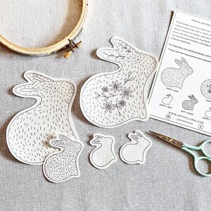 May include: Embroidery kit featuring pre-printed bunny designs. Includes a wooden embroidery hoop, scissors, and a guide with instructions. The bunny designs vary in size, with the largest measuring approximately 10cm x 11cm.