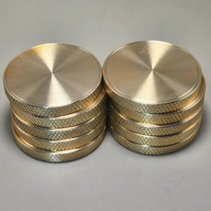 May include: Two stacks of gold-colored, circular metal objects. Each stack has a textured, diamond-patterned edge and a smooth, brushed top. The objects appear to be coasters or small decorative items.