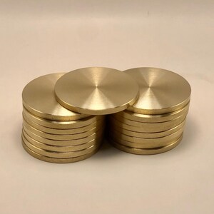 Puede incluir: Varias pilas de discos metálicos dorados. Cada disco es circular con una zona central elevada. Los discos están dispuestos en pilas ordenadas, con algunos discos colocados encima. Los discos parecen ser de latón.
