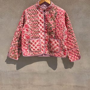 May include: A quilted jacket featuring a pink floral pattern with green leaves and a diamond-stitched texture. The jacket has a mandarin collar, long sleeves, two front patch pockets, and a pink and white striped trim along the edges and cuffs.