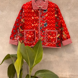 Puede incluir: Una chaqueta acolchada de color rojo vibrante con un estampado floral y una textura de costuras en forma de diamante. La prenda incluye cuello, mangas largas con puños a rayas y dos bolsillos delanteros, todo con ribetes a rayas rojas y blancas.