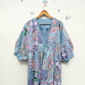 Könnte beinhalten: Ein Patchwork-Kleid in Blau-, Rosa- und Lilatönen mit floralen und geometrischen Mustern. Das Kleid hat einen V-Ausschnitt, Dreiviertelärmel mit gestreiften Manschetten und eine lockere, fließende Silhouette. Das Kleid hängt an einem Holzbügel.