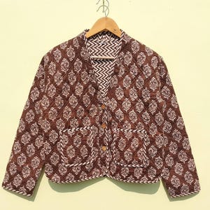 Peut inclure: Une veste matelassée marron avec un motif floral blanc. La veste a une fermeture boutonnée et deux poches.