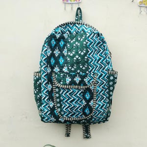 May include: A quilted backpack in shades of green, teal, and white, featuring a geometric pattern. The backpack has two front pockets and adjustable straps. The bag is hanging on a light-colored wall.