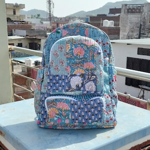 Puede incluir: Mochila acolchada con un estampado floral en tonos azules, rosas y blancos. La mochila tiene un compartimento principal, un bolsillo frontal y correas ajustables. La mochila está sobre una superficie azul claro.