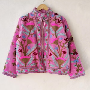 Peut inclure: Veste rose brodée de motifs floraux. La veste a un col montant et une fermeture éclair. Les broderies sont de couleurs marron, rose, bleu et vert. La veste est suspendue à un cintre en bois sur un mur blanc.