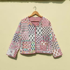 Puede incluir: Una chaqueta acolchada blanca con un diseño de patchwork que presenta varios patrones florales en rosa, azul y verde. La chaqueta tiene un cierre de botón y un ribete rojo alrededor de los bordes.
