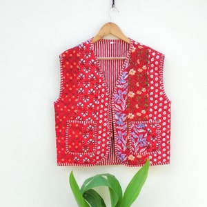 May include: A red quilted vest with a floral and polka dot pattern, hanging on a wooden hanger. The vest has a striped interior and two front pockets. The stitching is white, and the vest is displayed against a white background.