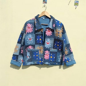 May include: A blue denim jacket with a patchwork design featuring embroidered flowers in pink, blue, and yellow. The jacket has a collared neckline and long sleeves.