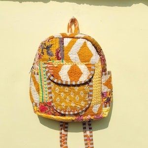 Puede incluir: Una mochila de patchwork amarilla, blanca y rosa con un pequeño bolsillo en la parte delantera. La mochila tiene un diseño acolchado y está hecha de tela de algodón.