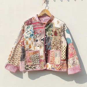 Pode incluir: Uma jaqueta acolchoada de patchwork com gola mandarim e fecho de botões. A jaqueta é feita de vários tecidos em tons de rosa, azul, marrom e branco, com padrões florais e geométricos.