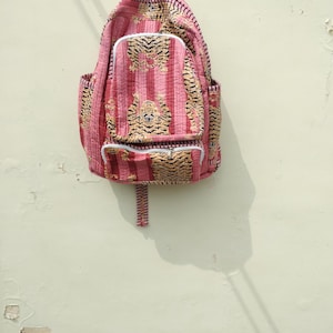 May include: A pink backpack with a tiger stripe pattern. The backpack has a front pocket with a zipper and two side pockets. The straps and handle are also pink. The backpack is hanging on a light-colored wall.