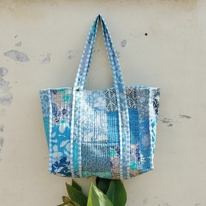 May include: A patchwork tote bag in shades of blue, featuring floral and geometric designs. The bag has handles with a blue and white gingham pattern. It hangs against a textured, off-white wall, with green leaves at the bottom.
