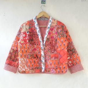 Pode incluir: Uma jaqueta acolchoada de patchwork em tons de laranja e rosa com padrões florais. A jaqueta tem um decote em V, mangas compridas e dois bolsos. O tecido é acolchoado e tem uma bainha branca.