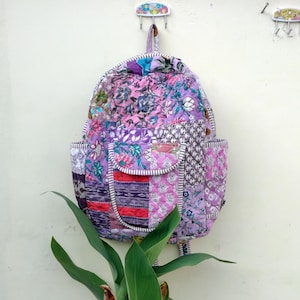 May include: A quilted backpack with a patchwork design in shades of purple, pink, and white. The backpack features floral patterns and a front pocket. It has two side pockets and a top handle for carrying.