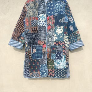 Pode incluir: Uma jaqueta patchwork em tons de azul, vermelho e branco, com uma variedade de padrões florais e geométricos. A jaqueta tem gola mandarim, mangas três quartos e forro listrado nos punhos e gola. A jaqueta está pendurada em um cabide de madeira.