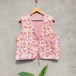 Pode incluir: Um colete branco sem mangas com estampa floral vermelha e laranja. O colete tem um decote em V e um fecho de nó na parte inferior.