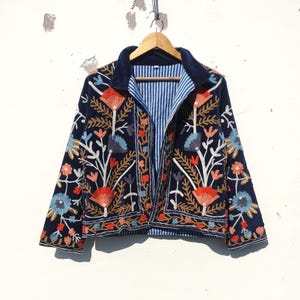 Peut inclure: Veste bleu marine ornée de broderies florales aux tons orange, rouge, bleu et blanc. La veste est dotée d'un col en velours bleu et d'une doublure rayée bleue et blanche. Elle est suspendue à un cintre en bois.