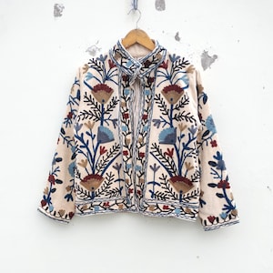 Könnte beinhalten: Beige Jacke mit aufwendiger Blumenstickerei in Blau-, Rot- und Brauntönen. Die Jacke hat einen hohen Kragen und hängt an einem Holzbügel. Die Stickerei bildet ein detailliertes Muster auf der Oberfläche der Jacke.