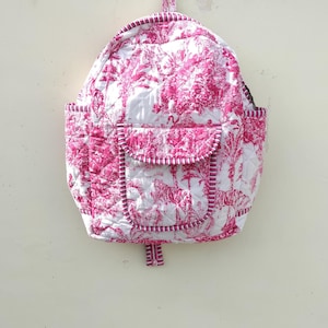 May include: A quilted backpack with a white background and a pink toile pattern featuring trees and animals. The backpack has a front pocket, side pockets, and a top handle. Pink and black striped trim outlines the edges.