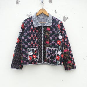 Puede incluir: Chaqueta acolchada con estampado floral en rojo, blanco y morado sobre fondo negro. La chaqueta tiene cuello a rayas y ribete de bolsillo. La chaqueta está colgada de una percha de madera.