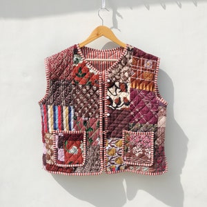 Pode incluir: Um colete acolchoado sem mangas com um design de patchwork em tons de vermelho, marrom e branco. O colete tem fecho de botão e dois bolsos.