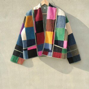 May include: A colourful patchwork corduroy jacket. The jacket is made up of squares of different colours, including blue, pink, green, and black. It is hung on a wooden hanger and has a striped lining and white stitching.