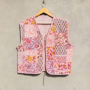 May include: A pink quilted vest with a patchwork design featuring floral and paisley patterns. The vest has a V-neck and a tie closure. The vest is hanging on a wooden hanger against a neutral background.