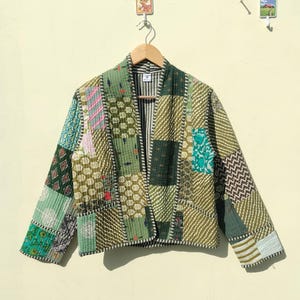 Pode incluir: Uma jaqueta patchwork em tons de verde, verde-azulado e creme pendurada em um cabide de madeira. A jaqueta apresenta uma variedade de padrões geométricos e florais, com uma textura acolchoada. A jaqueta tem um design sem gola e mangas compridas.
