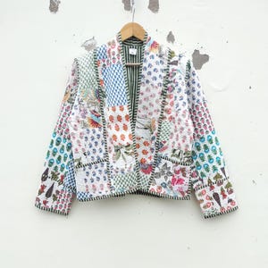 Puede incluir: Chaqueta patchwork con estampados florales y geométricos en tonos blancos, azules, rosas y verdes. La chaqueta tiene cuello y bolsillos, y está colgada de una percha de madera. La chaqueta está hecha de diferentes telas.