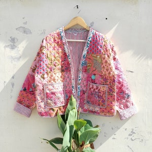 Pode incluir: Casaco acolchoado com design patchwork em tons de rosa e laranja, com estampas florais. O casaco tem forro listrado e dois bolsos frontais. Está pendurado em um cabide de madeira, medindo cerca de 70 cm de comprimento.