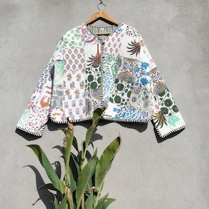 May include: A white quilted jacket with a patchwork design featuring various floral patterns in shades of blue, green, pink, and orange. The jacket has a zipper closure and a black trim.