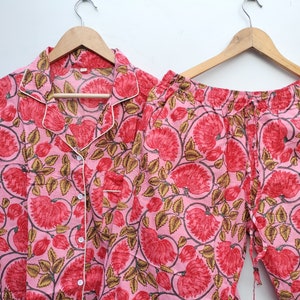 Puede incluir: Conjunto de pijama rosa con estampado floral con flores rojas y hojas verdes. La parte superior tiene cierre de botones y cuello. Los pantalones tienen cintura elástica y cordón.