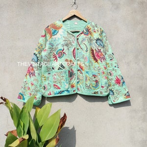 Pode incluir: Casaco acolchoado verde claro com um padrão floral em tons de rosa, azul e amarelo. O casaco tem dois bolsos frontais e está pendurado num cabide de madeira. O texto "THE VINTAGE ART CRAFTS" é visível no casaco.