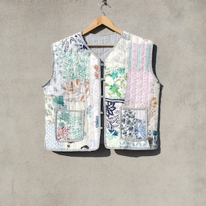 May include: A white quilted vest with a patchwork design featuring floral and geometric patterns in shades of green, blue, and pink. The vest has a zipper closure and two pockets.
