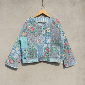 Pode incluir: Casaco patchwork com padrão floral em tons de azul, verde-azulado e rosa. O casaco é acolchoado e tem botões na frente e mangas compridas. O forro é de tecido listrado azul e branco. O casaco está pendurado em um cabide de madeira.