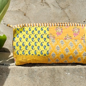 Puede incluir: Una bolsa de maquillaje acolchada amarilla con estampado paisley y floral. La bolsa tiene cierre de cremallera y un ribete a rayas marrón y blanco.
