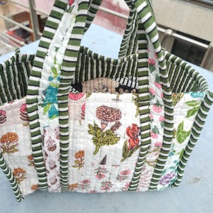 May include: A patchwork tote bag with a floral design. The bag has a white base with floral patterns in pink, orange, and green. The handles and trim are green and white striped. The bag is held up by a hand.