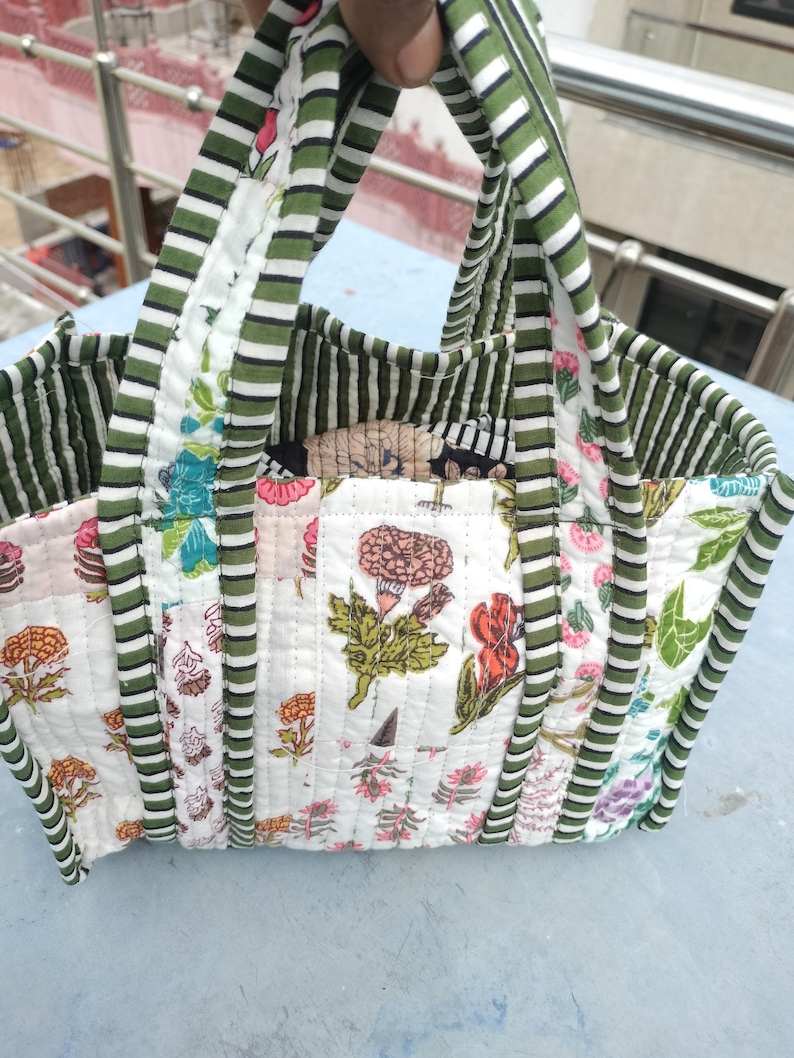May include: A patchwork tote bag with a floral design. The bag has a white base with floral patterns in pink, orange, and green. The handles and trim are green and white striped. The bag is held up by a hand.