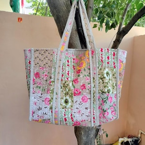 May include: A large, quilted tote bag with floral patterns in pink, yellow, and green on a beige background. The bag has long, striped handles and a rectangular shape, perfect for carrying items. The bag is hanging from a tree branch.