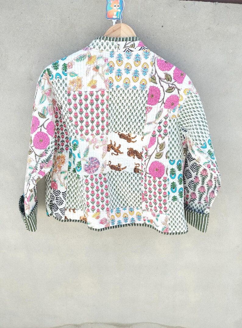 May include: A white quilted jacket with a patchwork design featuring floral and tiger motifs. The jacket has pink flowers, green leaves, and a striped cuff. The overall style is bohemian.