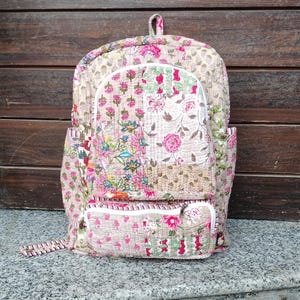 Puede incluir: Una mochila acolchada con un estampado floral en rosa, verde y beige. La mochila tiene un bolsillo frontal, bolsillos laterales y un asa superior. El diseño presenta un patchwork de estampados florales y una cremallera blanca.