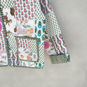 May include: A quilted jacket with a patchwork design featuring floral and animal motifs. The jacket has a white background with pink, green, and blue accents. The sleeves have a striped cuff. The jacket has a button closure and two pockets.